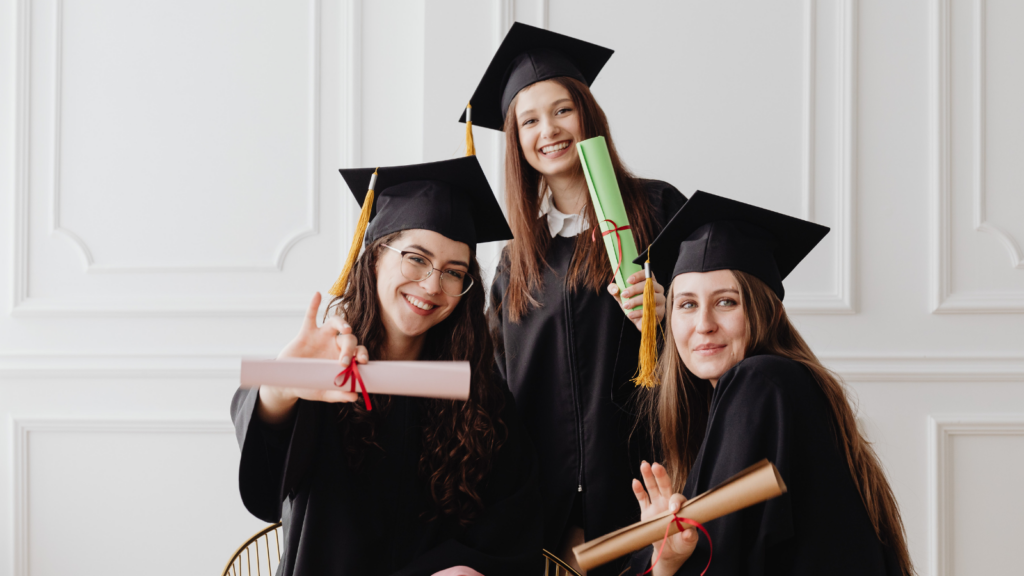 “Three happy female graduates in black caps and gowns holding diplomas and celebrating graduation together”